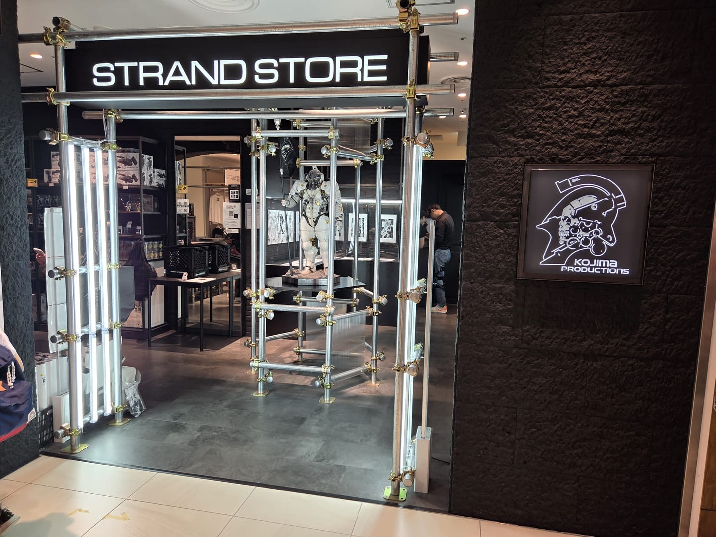 入り口がもうかっこいい。 単管パイプで組まれた入場口の上部に STRAND STORE と書かれた看板がある。入り口の右には KOJIMA PRODUCTIONS のロゴが飾られている。