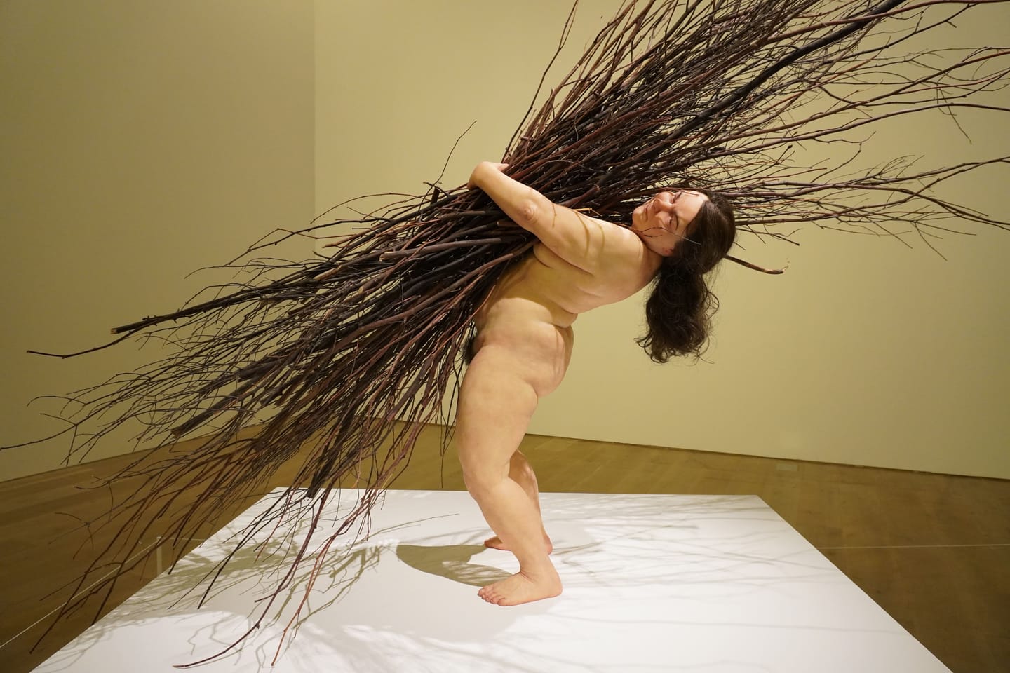 大量の枝を持って何とか持ちこたえている女性。 裸の女性が大量の木の枝を腕いっぱいに抱え込んで倒れないように耐えている。